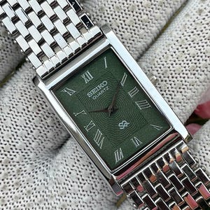 May include: A rectangular Seiko Quartz watch with a silver-tone metal link bracelet and a textured green dial. The watch face features silver Roman numeral hour markers, silver hands, and a central wave-like pattern. The Seiko logo is printed in white.