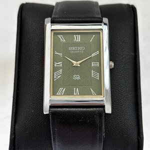 Op de afbeelding: Een rechthoekig Seiko Quartz horloge met een zilverkleurige kast en een zwarte leren band. De wijzerplaat van het horloge is olijfgroen met gouden Romeinse cijfers en wijzers. Het horloge wordt getoond op een zwart kussen.