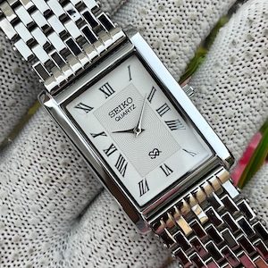 May include: A rectangular Seiko Quartz wristwatch with a silver-tone metal link bracelet. The white dial features black Roman numeral hour markers, a central textured wave pattern, and the text SEIKO QUARTZ printed in black near the top.
