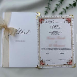 Könnte beinhalten: Eine weiße Nikkah-Heiratsurkunde-Mappe mit goldenem Schleifenband. Das innere Dokument zeigt verzierte florale Ränder, goldene Ecken und gedruckten Text auf Englisch und Arabisch, einschließlich des Satzes And We created you in pairs.