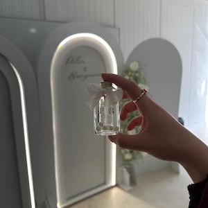 May include: A small, clear glass bottle with a cork stopper, adorned with a white ribbon bow at the neck. The front of the bottle features the black printed letters P and B separated by a vertical line, with several small white beads inside.