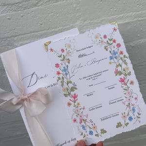 Könnte beinhalten: Eine weiße religiöse Heiratsurkunde mit Büttenrand, verziert mit einer bunten Blumenbordüre aus rosa, blauen und gelben Blüten. Das Dokument enthält türkischen Text, ein hellrosa Band auf der linken Seite und goldfarbene Metallecken als Akzente.