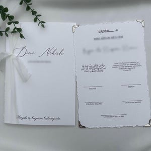Peut inclure: Un ensemble de certificat Dini Nikah blanc avec des bords effilochés et des accents d'angle argentés. La pièce de gauche présente un ruban blanc et le texte Dini Nikah et Hayirli es, huzurun baslangicidir. La droite inclut des lignes de signature.