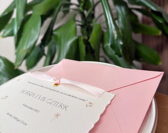 May include: A cream-coloured birth announcement card with scalloped edges, featuring the name SERRA EVIN OZTURK, date 8 december 2025, and stats 3460 g and 52 cm. The card is adorned with small floral illustrations and a pink ribbon, tucked into a pink envelope.