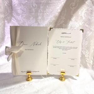 Könnte beinhalten: Ein weißes Nikkah-Zertifikatsset bestehend aus einer Mappe mit cremefarbener Schleife und einer passenden Karte mit Büttenrand, goldenen Ecken und schwarzem Text inklusive der Namen Elif und Ferhat, präsentiert auf goldenen Ständern.