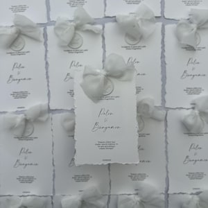 Op de afbeelding: Een verzameling rechthoekige witte papieren kaarten met geschepte randen, elk voorzien van een witte chiffon strik, een cirkelvormig waszegel en zwarte gedrukte tekst met de namen Pelin ve Bünyamin en een Turks citaat.