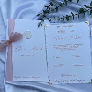 Könnte beinhalten: Ein weißes Dini Nikah-Zertifikatsset mit einem rosa Band, einem Wachssiegel und goldenen Ecken. Der Text enthält die Namen Selin & Furkan, religiöse Verse und Zeilen für Unterschriften, gedruckt auf strukturiertem Papier mit Büttenrand.