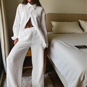 May include: White pajama set featuring a button-down shirt with a collar and a pair of wide-leg pants. The fabric appears to be lightweight and textured. The set is displayed in a bedroom setting.