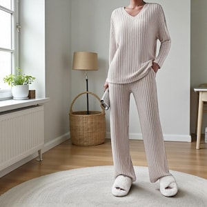 May include: A light beige ribbed knit pajama set with a V-neck top and wide-leg pants. The person is wearing fluffy white slippers. The set is made of a soft, comfortable material. The person is holding a small, folded, light-colored item.