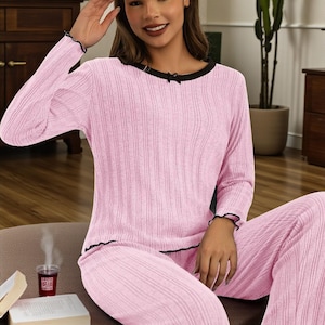 May include: Pink ribbed knit pyjama set with black trim. The long-sleeved top has a small bow at the neckline. The matching trousers complete the cosy sleepwear ensemble. The set is made of a soft, comfortable material.