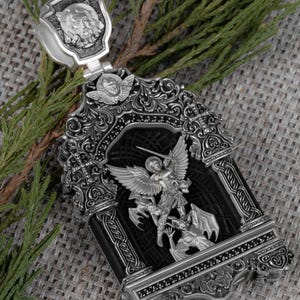 May include: Ornate silver pendant featuring a detailed depiction of Saint Michael slaying a dragon. The pendant has a rectangular black center, intricate scrollwork, and a shield-shaped bail with a carved face. The pendant is displayed on a textured gray surface.