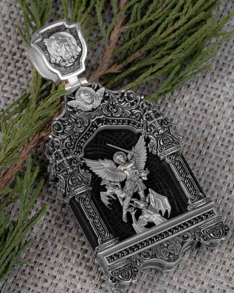 May include: Ornate silver pendant featuring a detailed depiction of Saint Michael slaying a dragon. The pendant has a rectangular black center, intricate scrollwork, and a shield-shaped bail with a carved face. The pendant is displayed on a textured gray surface.