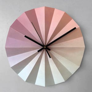 May include: A round wall clock with a fan-like design, featuring a gradient of pink, peach, and white hues. The clock has black hands and a gold center, creating a modern and artistic aesthetic. The clock's diameter is approximately 12 inches.
