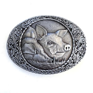 Puede incluir: Una hebilla de cinturón ovalada de metal plateado con un diseño en relieve de dos cerdos descansando en la hierba. La imagen central de los cerdos está rodeada por un borde ornamentado de motivos florales y de enredaderas con un borde de cuerda.