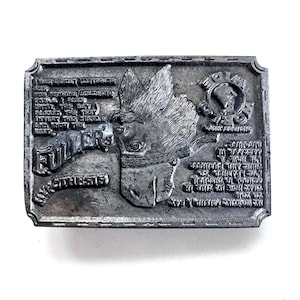 Peut inclure: Une boucle de ceinture rectangulaire en métal argenté présentant un relief d'une tête de cheval au centre. La surface est gravée avec le texte "FULLER'S HYPOTHESIS" et "WIDE LOAD" ainsi que plusieurs lignes de petites lettres stylisées.