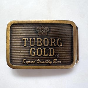 May include: A rectangular brass belt buckle with the words "TUBORG GOLD" and a crown emblem. The buckle also features the text "Export Quality Beer" and has a textured, ridged background.