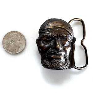 May include: A bronze-toned metal belt buckle featuring a sculpted relief of a pirate head wearing a bandana and an eye patch, positioned next to a United States quarter coin for scale, showing the detailed facial features and the buckle loop.