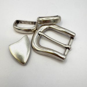Puede incluir: Un conjunto de componentes de hebilla de cinturón de metal plateado, que incluye una hebilla, una trabilla y una pieza decorativa en forma de escudo. Las piezas tienen un aspecto ligeramente envejecido.