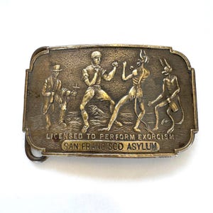 May include: A vintage metal belt buckle with a detailed relief scene. The buckle features figures engaged in a boxing match with demons, and a man holding a cross. The text "LICENSED TO PERFORM EXORCISM" and "SAN FRANCISCO ASYLUM" are inscribed.