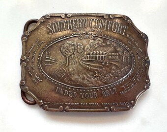 May include: A rectangular brass belt buckle with rounded edges featuring an embossed scene of a riverboat and a house. The text reads SOUTHERN COMFORT, THE GRAND OLD DRINK OF THE SOUTH, UNDER YOUR BELT, and NOT GENUINE WITHOUT THIS STEEL ENGRAVED LABEL.