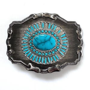May include: A rectangular silver-toned belt buckle with a brushed metal finish and ornate, scrolled edges. The center features a large, oval turquoise stone surrounded by a double border of small, round and elongated turquoise cabochons.