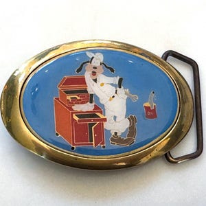 Pode incluir: Fivela de cinto oval com moldura dourada e centro azul. A imagem apresenta o Pateta com roupa de mecânico, com uma caixa de ferramentas, lata de óleo e chave inglesa. Artigo vintage.
