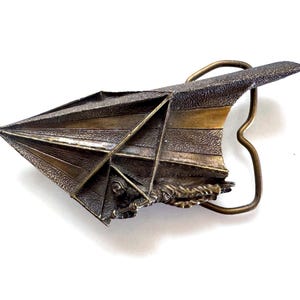 May include: A vintage metal belt buckle with a detailed design. The buckle features a textured, bronze-colored airplane or glider shape with a brass-colored frame. The design includes lines and geometric shapes, suggesting a historical or artistic theme.