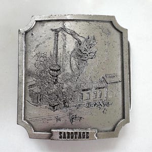 Könnte beinhalten: Rechteckige, silberfarbene Metallschnalle mit einer gravierten Industrieszene. Das Wort "SABOTAGE" ist unten abgebildet. Die Schnalle zeigt eine detaillierte Darstellung einer Fabrik und eines Krans.