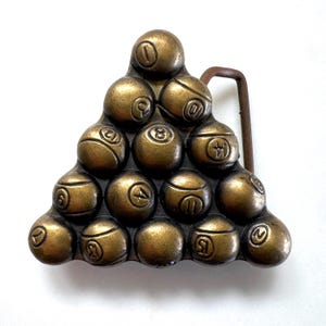 Puede incluir: Una hebilla de cinturón de metal color latón con forma de triángulo de billar. El diseño presenta quince esferas individuales, cada una grabada con un número único, dispuestas en forma de pirámide con un lazo de metal en el lado derecho.