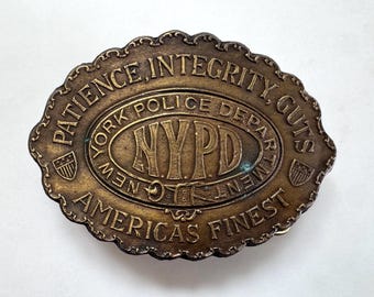 Puede incluir: Una hebilla de cinturón ovalada de latón con borde festoneado que presenta el texto en relieve PATIENCE, INTEGRITY, GUTS, NEW YORK POLICE DEPARTMENT, NYPD y AMERICAS FINEST. Dos pequeños iconos de escudo están en los lados.