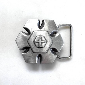 May include: A silver-toned metal belt buckle featuring a six-pointed star shape with deep notches between each point. The center of the buckle displays a raised hexagonal emblem with a cross design, and a rectangular loop is attached to the side.