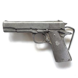 Puede incluir: Una hebilla de cinturón de metal plateado con forma de pistola semiautomática. El diseño presenta una empuñadura texturizada, un guardamonte y un lazo de metal en la parte posterior para sujetarlo al cinturón. El acabado es de aspecto mate.
