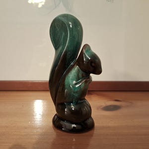 Puede incluir: Una figura de ardilla de cerámica con acabado brillante. La ardilla es principalmente verde oscuro y marrón, con una cola rizada y una pose sentada. La figura está sobre una superficie de madera.