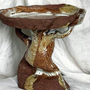 Può includere: Una ciotola in ceramica su piedistallo con una parte superiore circolare, ampia e piatta, sostenuta da una base scolpita. L'oggetto è realizzato in argilla testurizzata marrone rossiccio con accenti di smalto bianco e marrone chiaro.