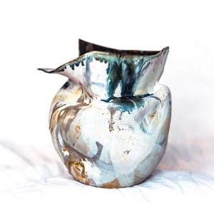 Pode incluir: Um vaso de cerâmica com uma forma orgânica irregular e uma borda larga e alargada. A superfície apresenta um esmalte marmoreado em tons de branco, azul profundo e castanho terroso, com gotas e redemoinhos criando um aspeto fluido e colorido.