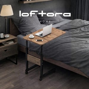 May include: A wooden laptop table with a black metal frame is positioned next to a bed with gray bedding. A laptop, a cup, and a phone are on the table. A wooden nightstand with a lamp and a drawer is on the left. The logo "loftora HOME" is in the image.