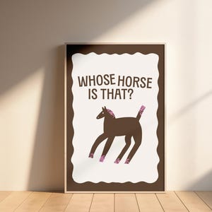 May include: A framed art print with a brown horse illustration and pink details. The text "WHOSE HORSE IS THAT?" is above the horse. The artwork is set against a cream-coloured background. The frame is brown and the print is on a wooden floor.