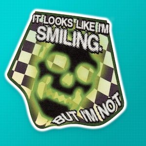 May include: A patch with the text "IT LOOKS LIKE I'M SMILING BUT I'M NOT" in pixelated white font. The patch features a green skull design with a checkered background in shades of green and black.