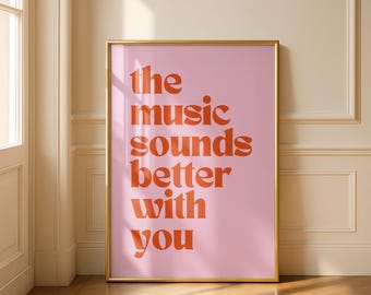 May include: A framed art print with the text "the music sounds better with you" in orange lettering on a pink background. The frame is gold. The artwork is displayed on a wooden floor.