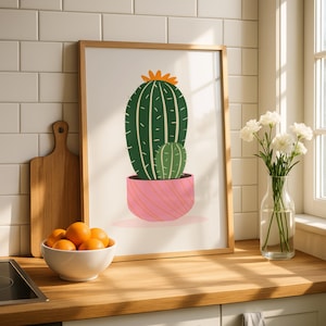 May include: A framed wall art print featuring a dark green cactus with a smaller light green cactus beside it, both potted in a pink planter with orange diagonal lines. The cactus is topped with a small orange flower on a white background.
