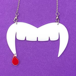 Puede incluir: Un collar de acrílico blanco con un diseño estilizado de colmillos de vampiro y una gota de sangre roja colgante. La cadena plateada cuelga sobre un fondo morado. Un accesorio único para Halloween o fiestas de disfraces.
