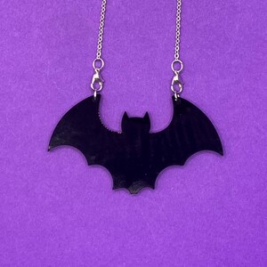 Può includere: Una collana con ciondolo a forma di pipistrello nero e catena argentata, su sfondo viola. Il ciondolo a pipistrello è una silhouette piena con ali spiegate. Un accessorio a tema Halloween.