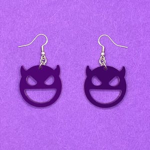 May include: A pair of purple devil face earrings with gold-coloured hooks. The earrings feature a smiling face design with horns. The earrings are set against a purple background.