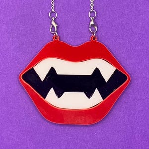 Puede incluir: Un collar con colgante de dientes de vampiro rojos, negros y blancos. El colgante tiene forma de boca con afilados dientes blancos y un interior negro. El collar cuelga de una cadena plateada sobre un fondo morado.