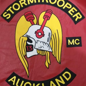 Puede incluir: Un parche de cuero rojo con la palabra "STORMTROOPER" en amarillo, arqueada en la parte superior. Un gráfico de casco con alas amarillas y detalles rojos está en el centro. El número "13" es visible. La palabra "AUCKLAND" está arqueada en la parte inferior.