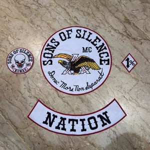 Op de afbeelding: Een verzameling geborduurde patches met de tekst "SONS OF SILENCE" en "NATION". De patches tonen een schedel, een adelaar en de tekst "Donec Mors Non Separat". De patches zijn wit met rode en zwarte accenten.