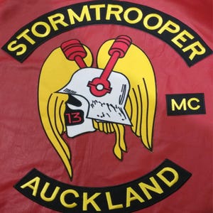 May include: A red jacket patch with the word "STORMTROOPER" in yellow arched text above a graphic of a white helmet with red and yellow accents. Below, the word "AUCKLAND" is arched in yellow. A small "MC" patch is on the right.