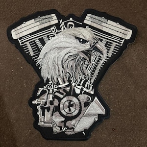 May include: A black and white embroidered patch featuring an eagle's head superimposed over a motorcycle engine design. The eagle is detailed with gray shading, and the engine components are rendered in a technical style.