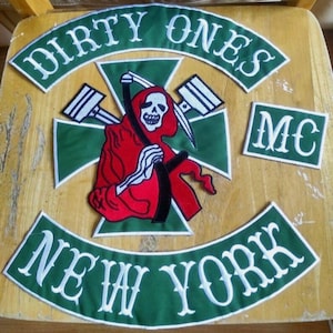 May include: Embroidered patches featuring the words "DIRTY ONES" and "NEW YORK" in white on green fabric. A central patch depicts a Grim Reaper figure in red, holding a scythe, with a skull face. The letters "MC" are also present.