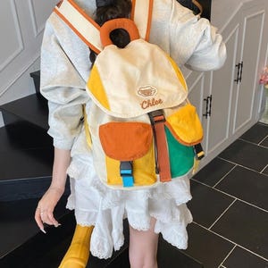 May include: A multi-coloured backpack featuring a cream-coloured top flap with the text Chlee embroidered in brown. The bag includes a bright orange front pocket, a yellow base, a green side pocket, and adjustable straps with blue and black buckles.